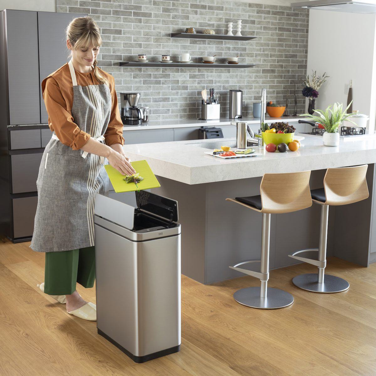Thùng rác cảm biến EKO đặt cạnh bàn đảo bếp, người dùng gạt rác sơ chế thẳng vào thùng